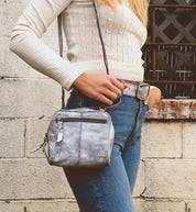 A person in blue jeans and a cream sweater stands against a textured stone wall, holding the Capture crossbody bag made of eco-friendly leather by Bed Stu.