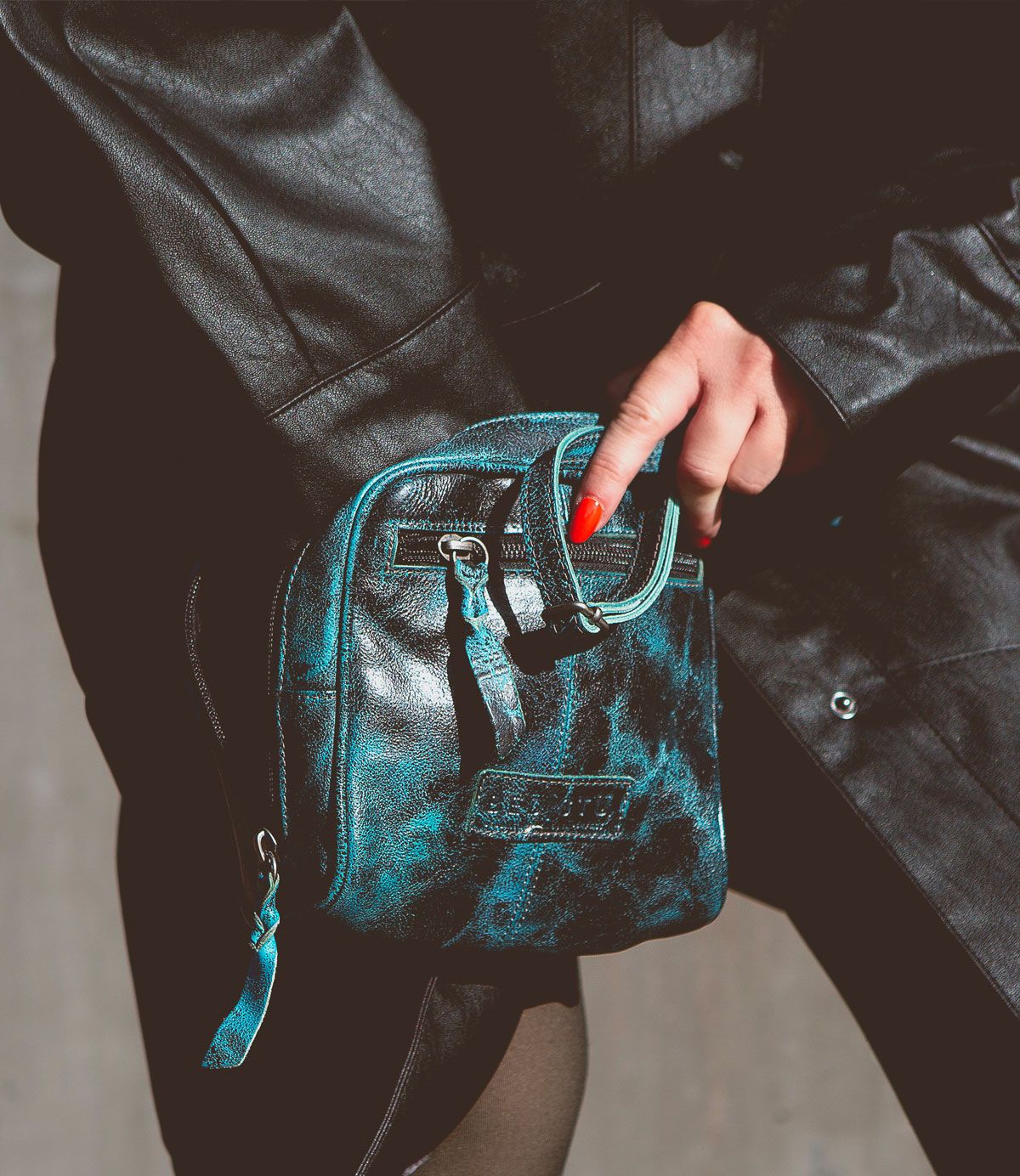 Person wearing a black leather jacket holding a small, artisan-made black and teal crossbody bag called Capture by Bed Stu.