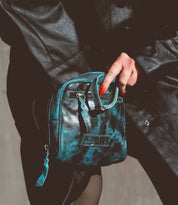 Person wearing a black leather jacket holding a small, artisan-made black and teal crossbody bag called Capture by Bed Stu.