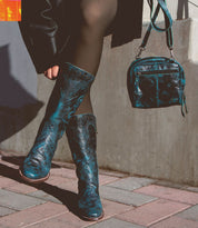 A person wearing blue patterned knee-high boots and black tights, holding a compact blue handbag from the "Capture" collection by Bed Stu with a matching pattern, is standing on a tiled sidewalk near a concrete wall. The handbag boasts eco-friendly leather, reflecting their commitment to sustainable fashion.