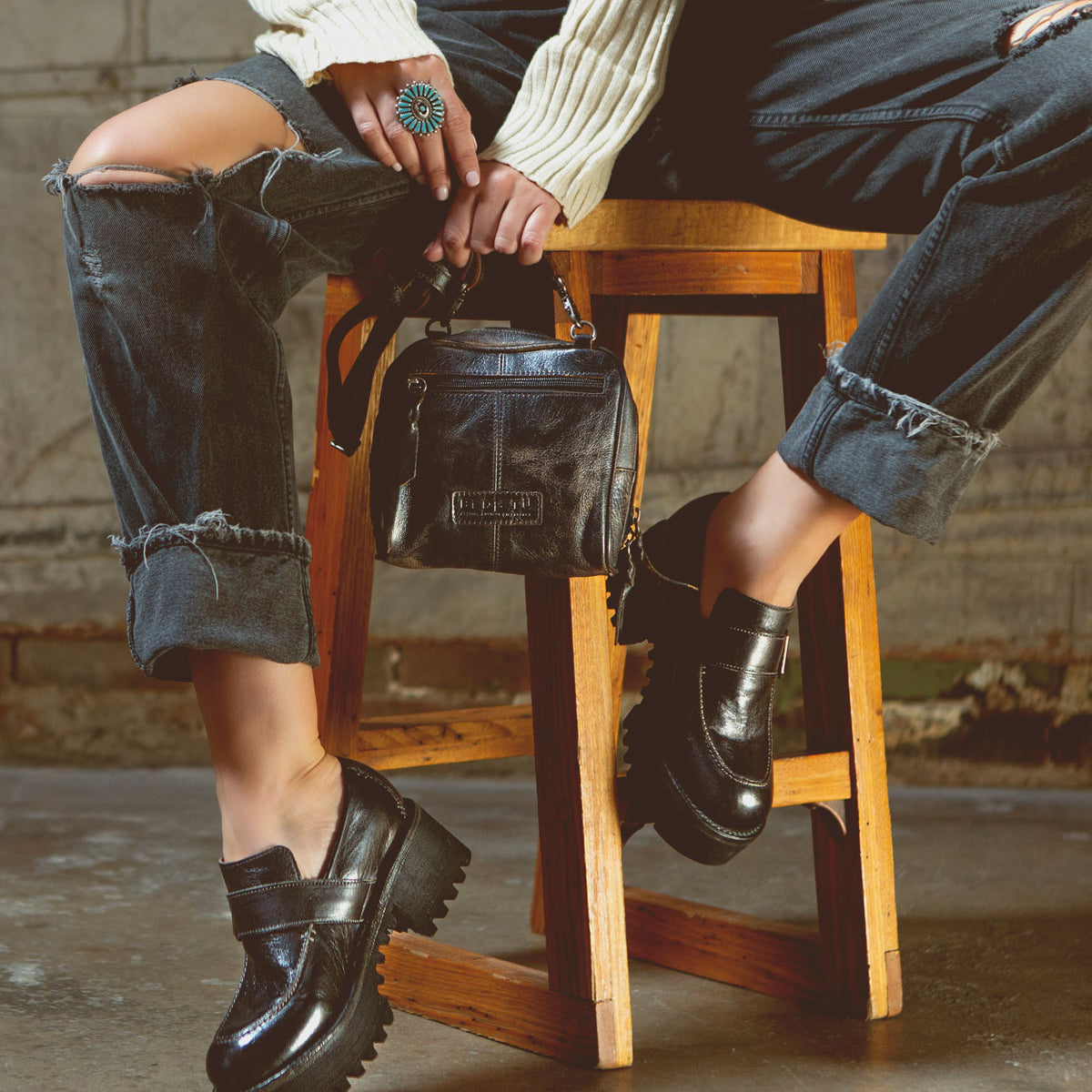 A petite woman is sitting on a stool elegantly holding a Bed Stu Capture eco-friendly leather purse, creating a captivating capture.