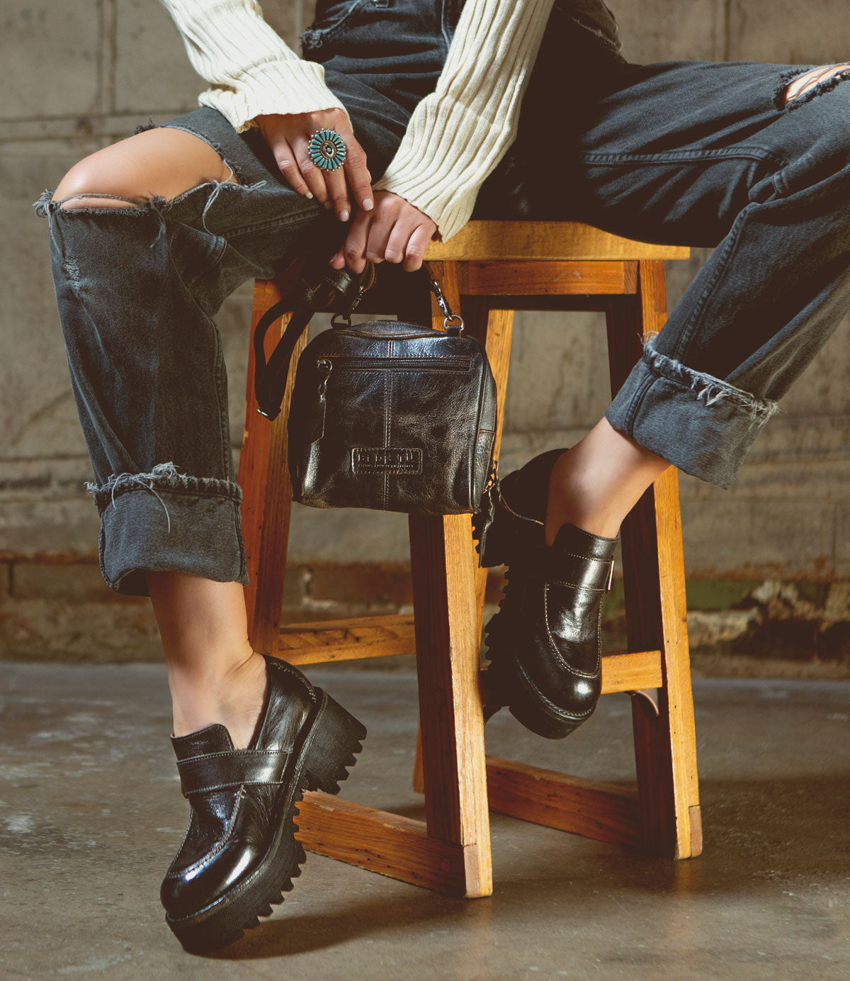 A petite woman is sitting on a stool elegantly holding a Bed Stu Capture eco-friendly leather purse, creating a captivating capture.