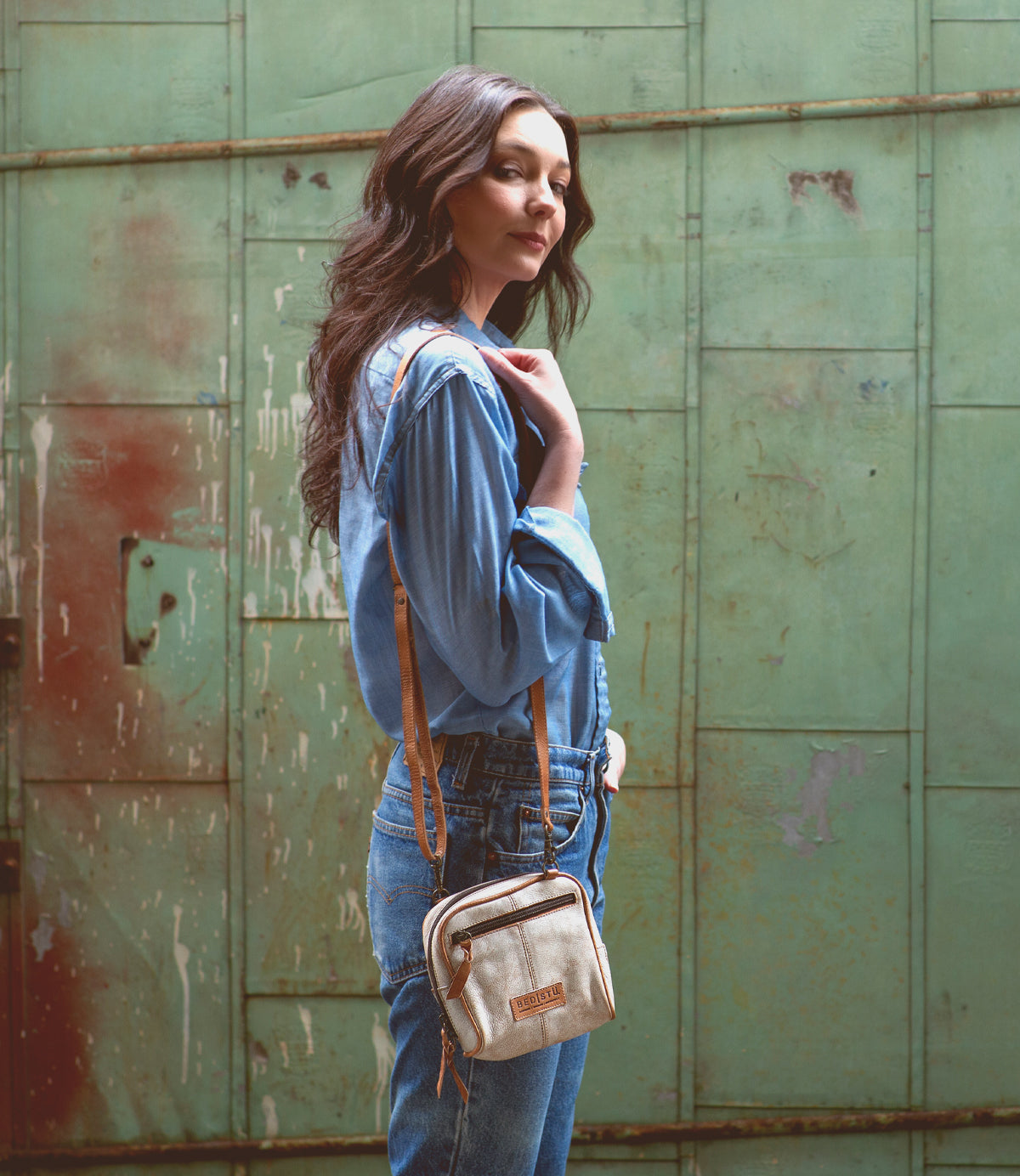 An eco-friendly woman in Capture denim overalls standing in front of a green wall.