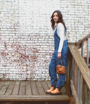 A woman wearing denim overalls capturing the eco-friendly spirit while standing on a wooden deck with an elegant and compact bag made of sustainable materials, the Bed Stu Capture showcases her commitment to sustainability.