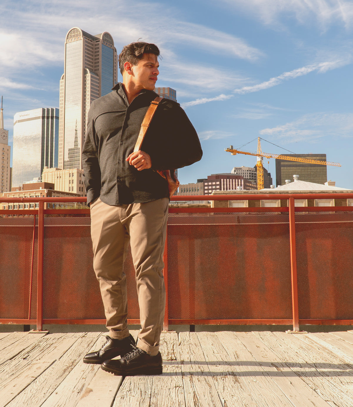 A man in a dark shirt and khaki pants stands on a wooden walkway, effortlessly stylish with his Barton leather high-top sneakers from Bed Stu. A shoulder bag rests at his side as he gazes at the skyline of skyscrapers and cranes on a bright, sunny day.
