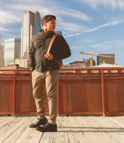 A man in a dark shirt and khaki pants stands on a wooden walkway, effortlessly stylish with his Barton leather high-top sneakers from Bed Stu. A shoulder bag rests at his side as he gazes at the skyline of skyscrapers and cranes on a bright, sunny day.