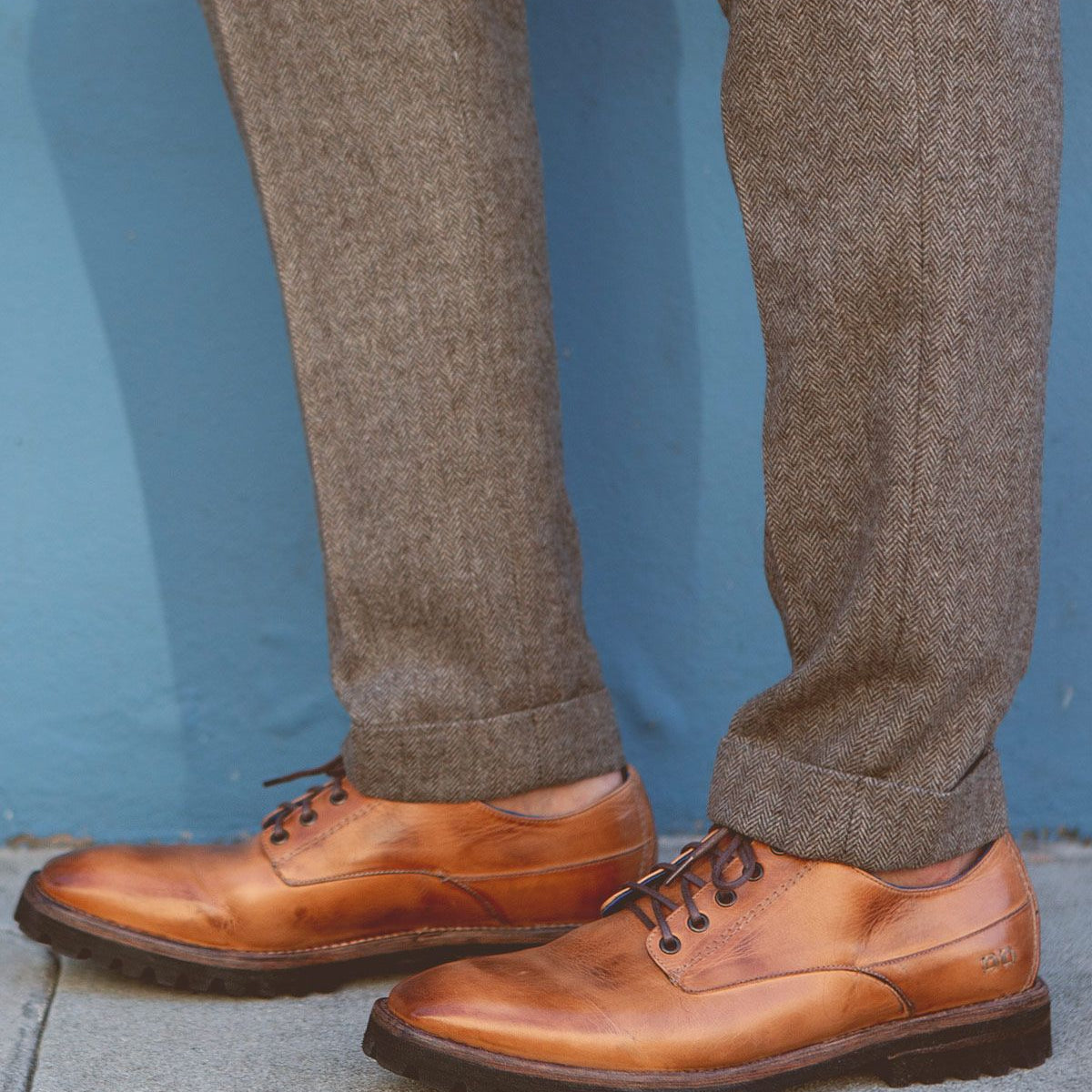A person wearing the Carter shoes by Bed Stu, made from vegetable-tanned leather, and gray herringbone trousers stands on a concrete surface against a blue wall.