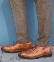A person wearing the Carter shoes by Bed Stu, made from vegetable-tanned leather, and gray herringbone trousers stands on a concrete surface against a blue wall.
