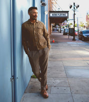 A man leans against a blue wall on the sidewalk, sporting a brown jacket and gray pants. His Carter shoes by Bed Stu, crafted with Goodyear welt construction, catch the eye. In the background, a "Now Playing" sign adds to the scene's charm.
