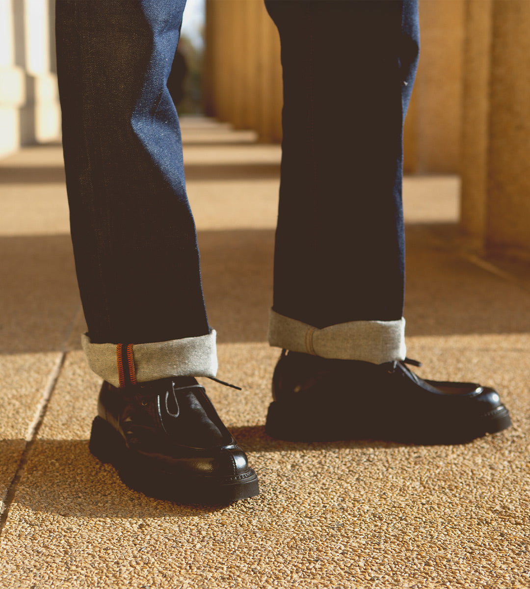 A person stands on textured ground wearing cuffed jeans and sleek Casotto moc-toe lace-up boots from Bed Stu, expertly crafted from Italian vegetable-tanned leather.