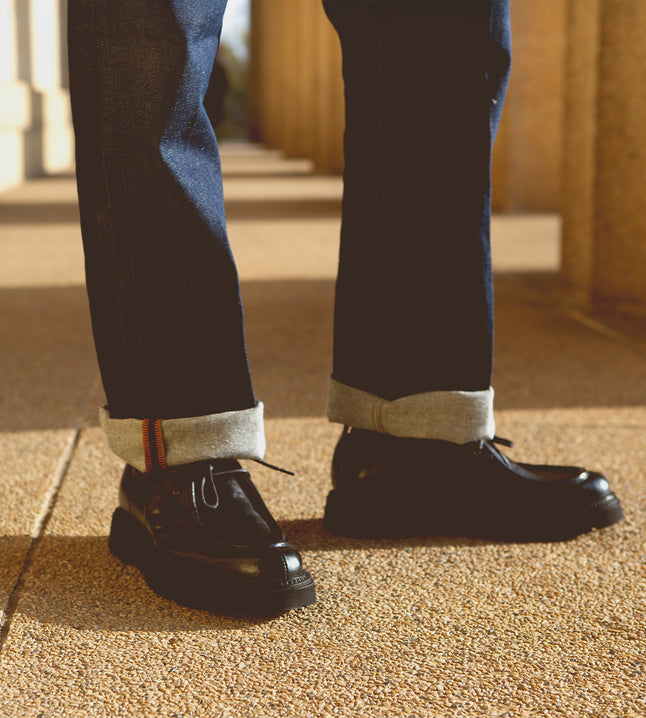 A person stands on textured ground wearing cuffed jeans and sleek Casotto moc-toe lace-up boots from Bed Stu, expertly crafted from Italian vegetable-tanned leather.
