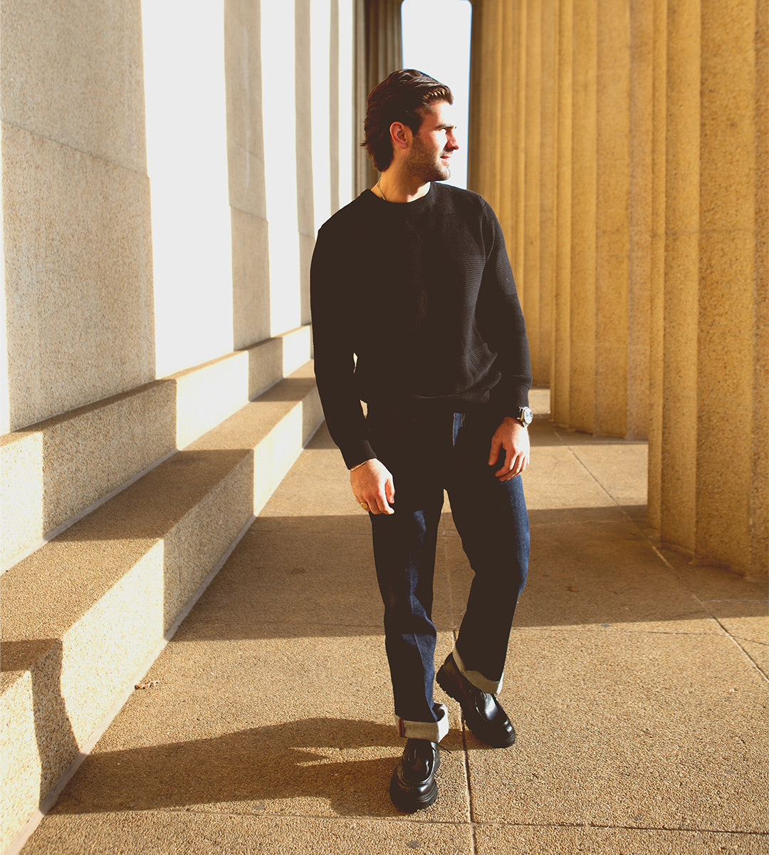 A person in a black sweater and jeans strolls between stone columns, their shadow stretching under the sunlight. The smooth stride is accentuated by Bed Stu's Casotto boots, which reflect exquisite Italian craftsmanship.