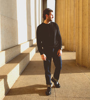 A person in a black sweater and jeans strolls between stone columns, their shadow stretching under the sunlight. The smooth stride is accentuated by Bed Stu's Casotto boots, which reflect exquisite Italian craftsmanship.