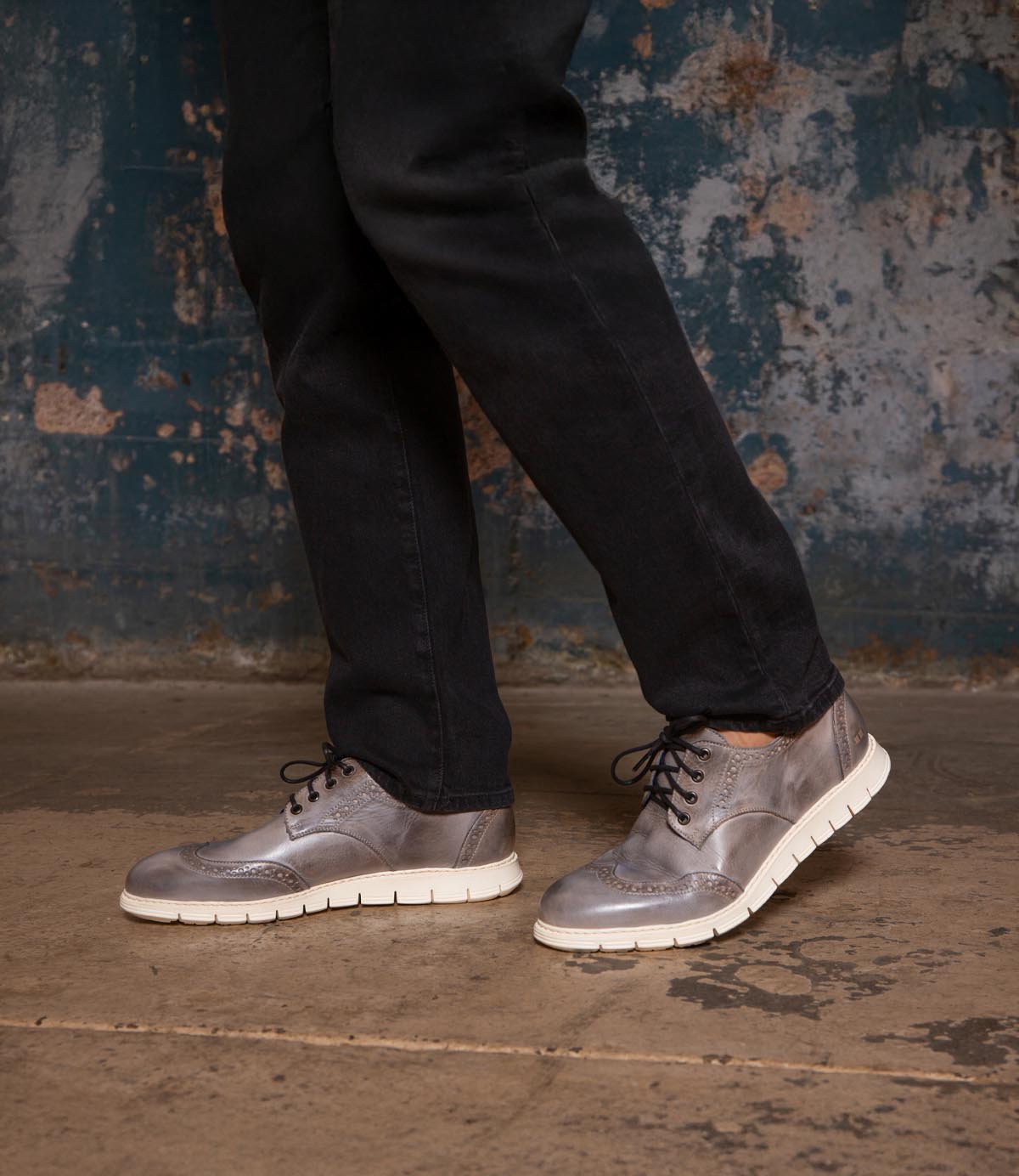 Someone wearing black pants and Bed Stu Cayuga II gray leather wingtip derby shoes with brogue details and white soles stands on a concrete floor against a distressed wall.