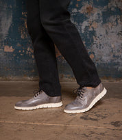 Someone wearing black pants and Bed Stu Cayuga II gray leather wingtip derby shoes with brogue details and white soles stands on a concrete floor against a distressed wall.