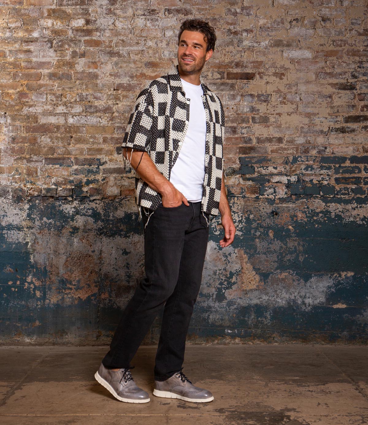 A man stands indoors by a brick wall, smiling and looking aside. He wears a patterned shirt, white tee, black pants, and Bed Stu Cayuga II casual men's shoes with brogue detailing.