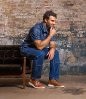 Wearing Bed Stu's Cayuga II casual brogue shoes, a man in a blue shirt and jeans sits smiling on a brown leather bench in front of a weathered brick wall.