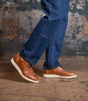 A person wearing blue jeans and Bed Stu Cayuga II brown leather wingtip derby shoes with brogue details and white soles stands on a concrete floor against a textured wall.
