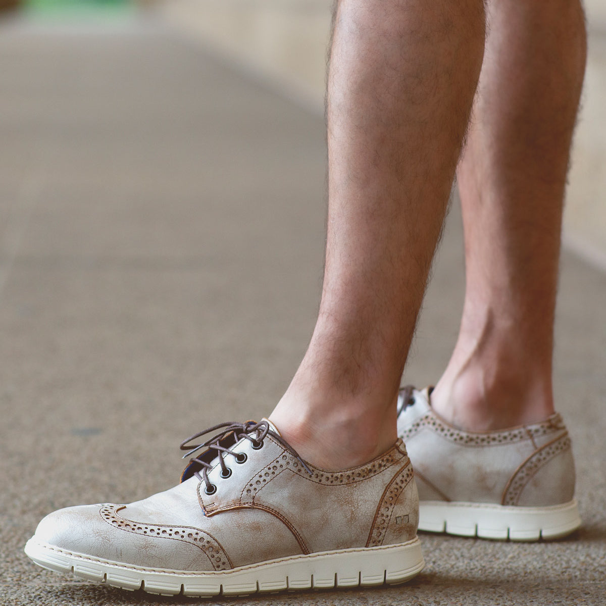 A casual male with bare legs wears Bed Stu Cayuga II lightweight leather wingtip shoes featuring brogue details and white soles, standing on a textured surface.
