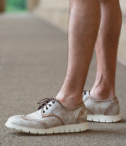 A casual male with bare legs wears Bed Stu Cayuga II lightweight leather wingtip shoes featuring brogue details and white soles, standing on a textured surface.