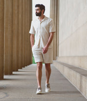 A casual male with short dark hair and a beard is walking along a corridor between stone columns, wearing a light-colored short-sleeved shirt, matching shorts, and lightweight Bed Stu Cayuga II leather wingtip shoes with brogue details, his hands resting comfortably in his pockets.