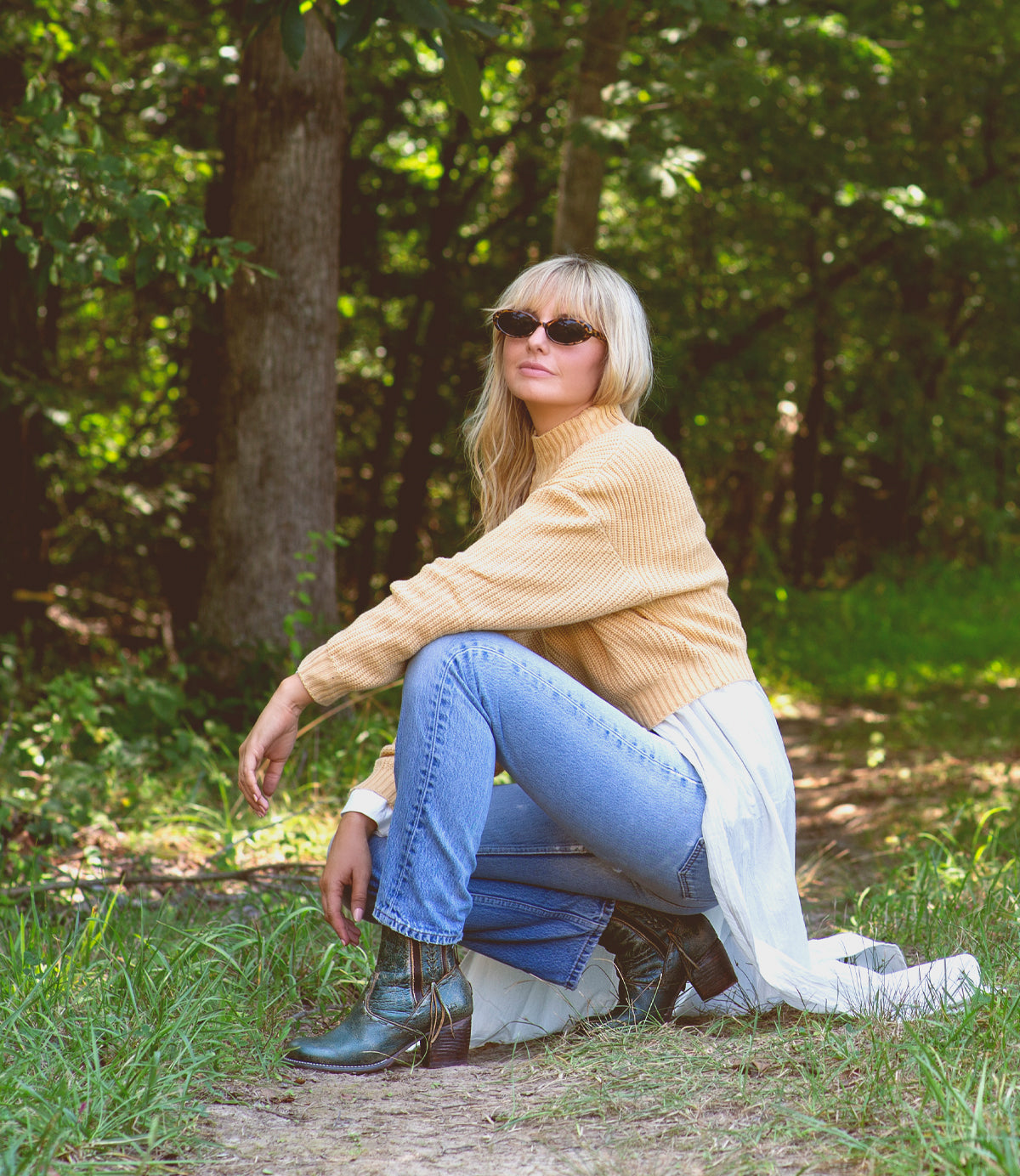 A woman crouching down in the woods wearing Bed Stu Celestine ankle boots and a yellow sweater.