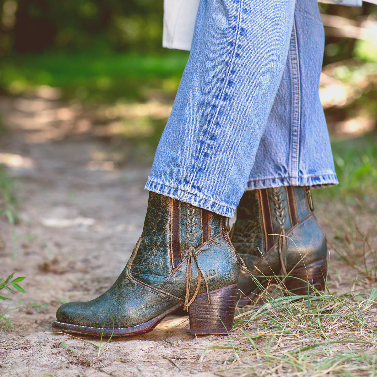 A woman wearing jeans and a pair of green Celestine western-inspired ankle boots from Bed Stu with hem stitch details.