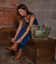 A woman in a denim jumpsuit ties brown lace-up boots while the Bed Stu Celindra LTC, a spacious zip-top tote, rests beside her—perfect for carrying everyday essentials.