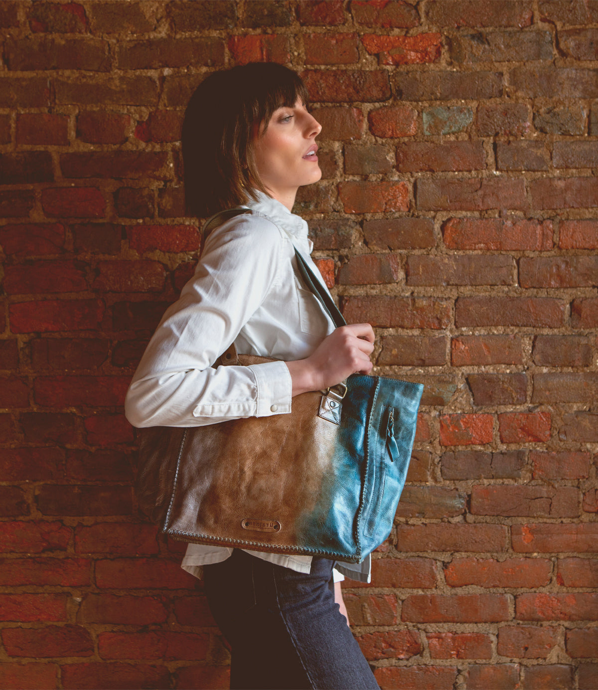 Bed Stu Celindra LTC, a woman, holding a zip-top tote purse against a brick wall.