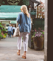 A woman with long hair, wearing a denim jacket, white pants, and sandals, carrying a spacious Bed Stu Celindra LTC with a secret pocket, walks down a path next to a garden center with plants and flowers.