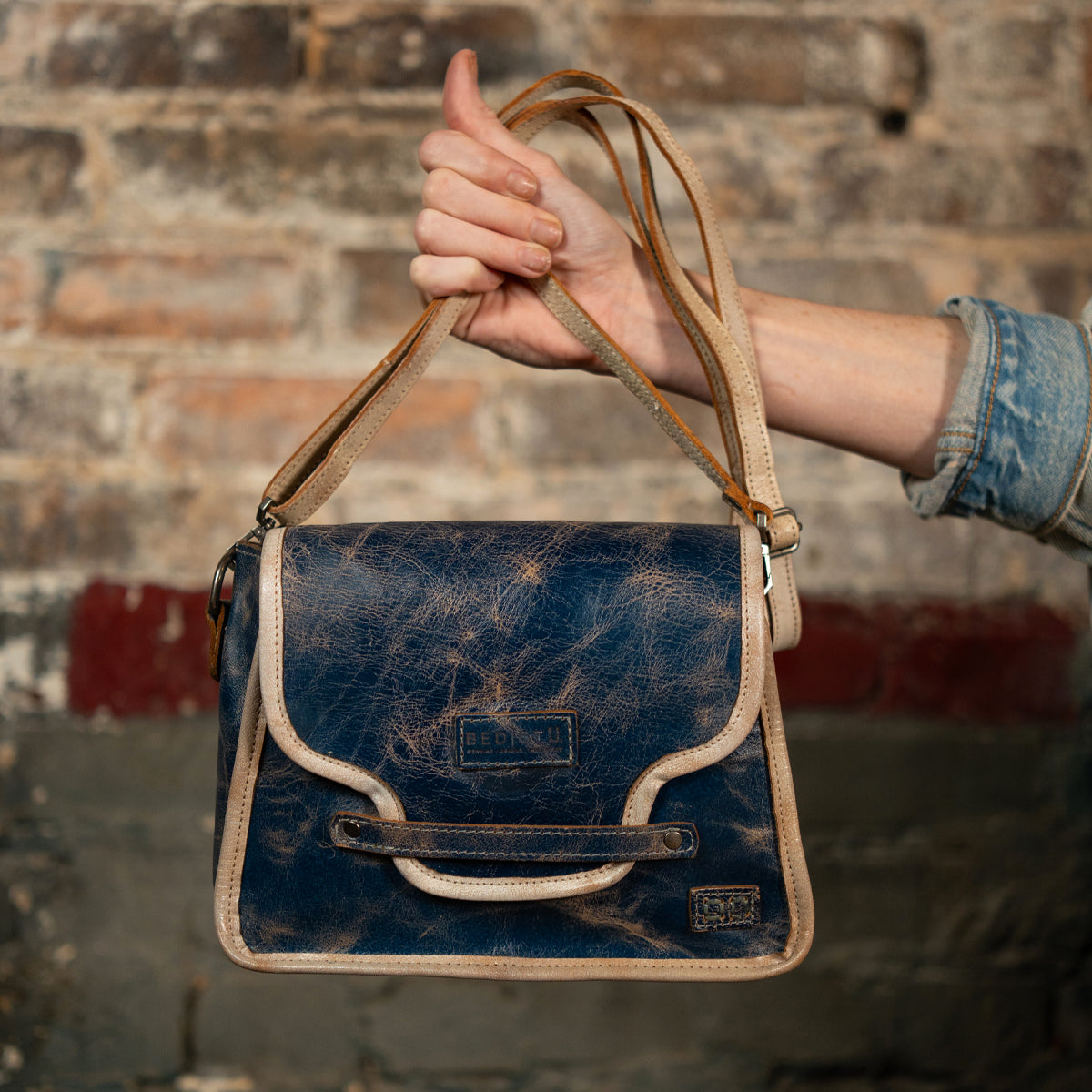 A person in a denim shirt holds up the Bed Stu Ceremonious, a vintage-style blue leather flap-top bag with tan trim and magnetic closure, against a rustic brick wall.