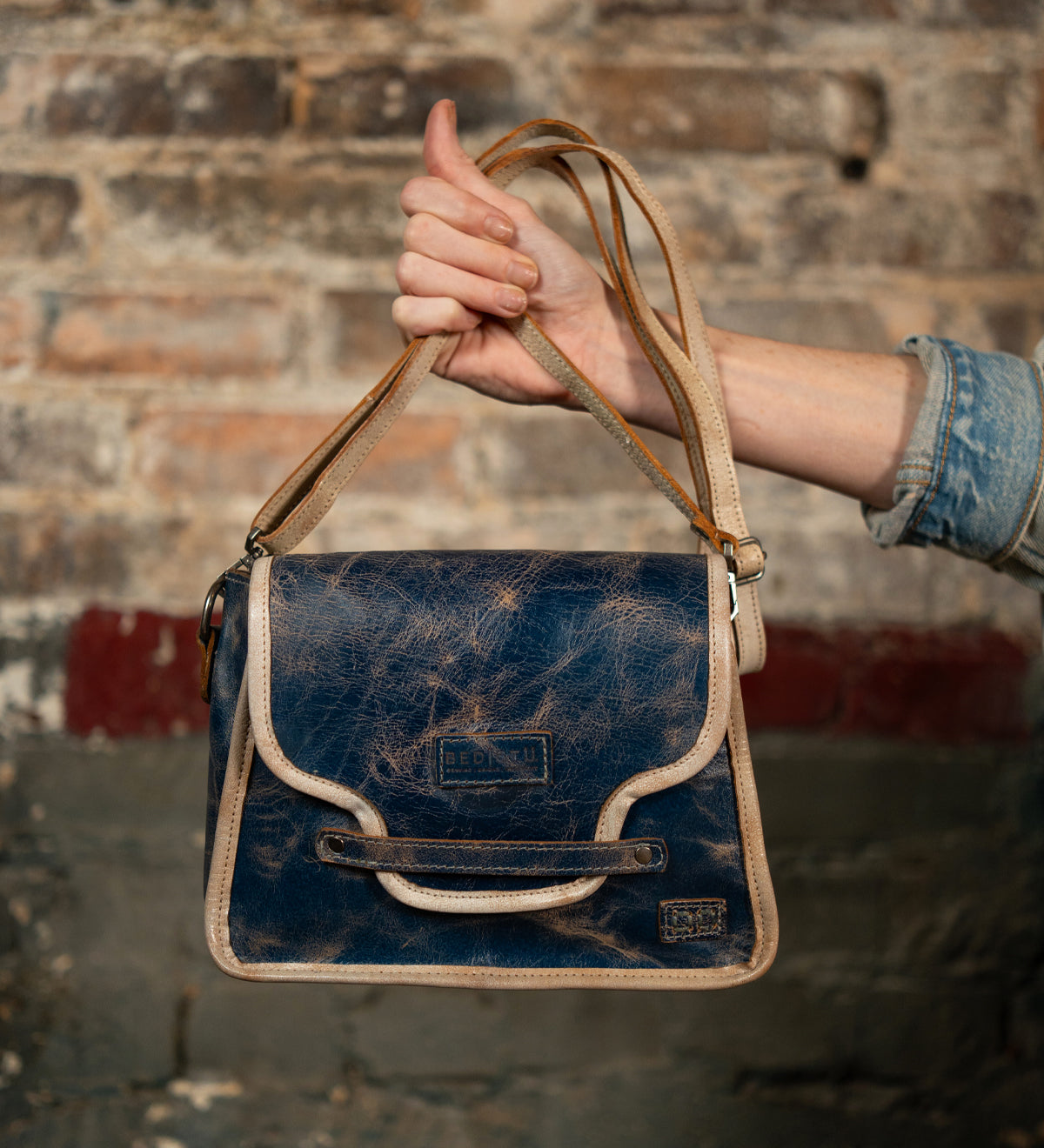 A person in a denim shirt holds up the Bed Stu Ceremonious, a vintage-style blue leather flap-top bag with tan trim and magnetic closure, against a rustic brick wall.