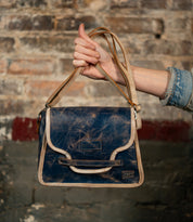 A person in a denim shirt holds up the Bed Stu Ceremonious, a vintage-style blue leather flap-top bag with tan trim and magnetic closure, against a rustic brick wall.