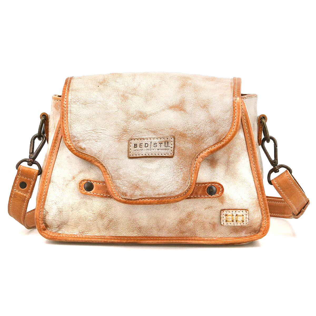 The Bed Stu Ceremonious is a beige and tan flap-top bag with a magnetic closure, metal clasps, embossed logo patch, and adjustable shoulder strap, shown against a white background.