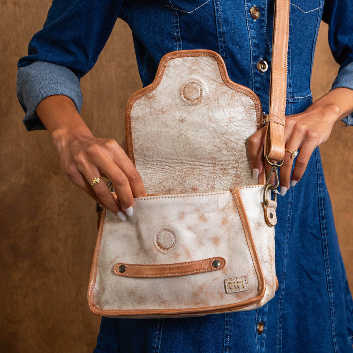 A person in a blue denim dress holds the open Ceremonious flap-top bag by Bed Stu, featuring light-colored leather with tan strap and trim, showing the inside pocket.