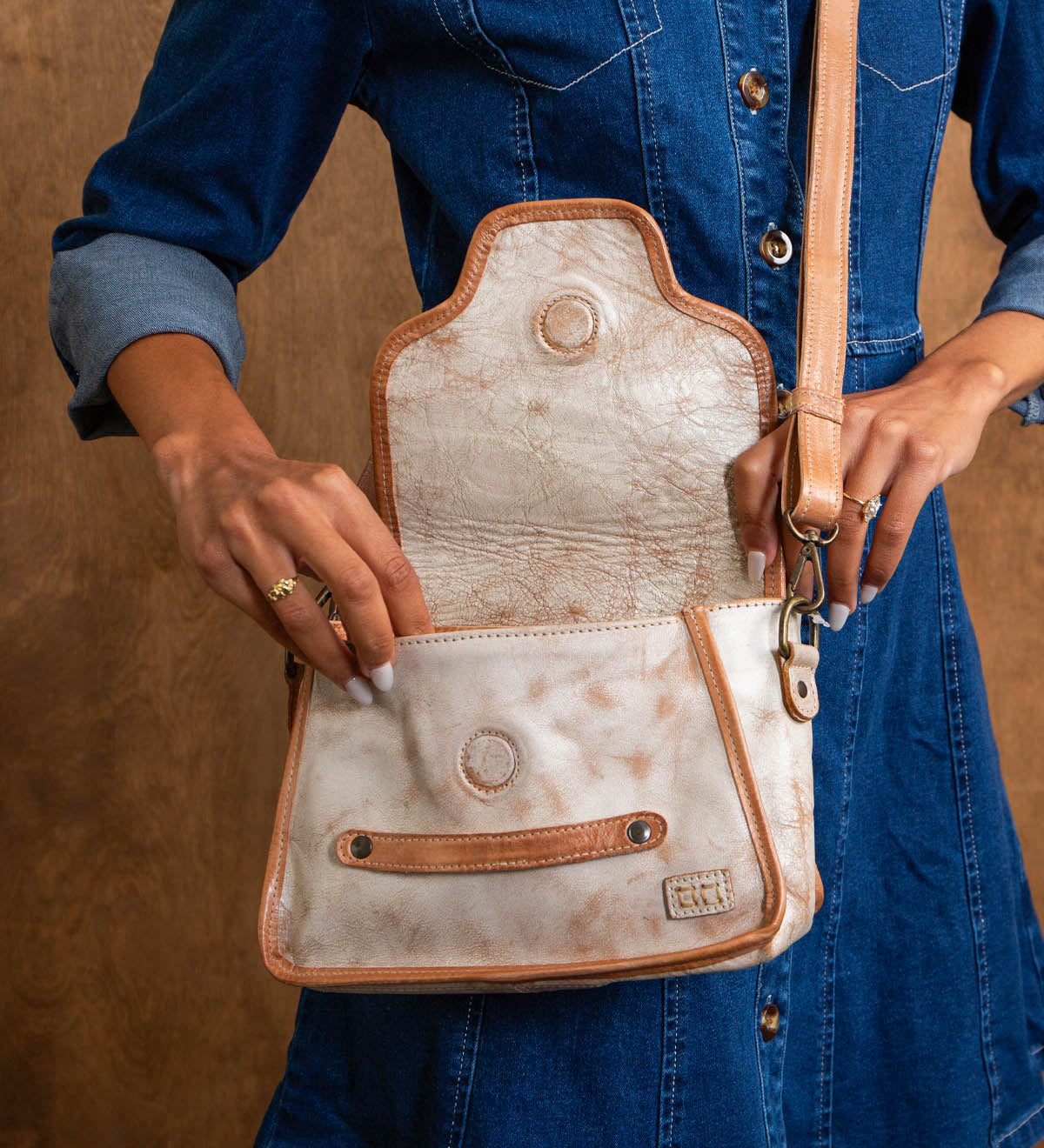 A person in a blue denim dress holds the open Ceremonious flap-top bag by Bed Stu, featuring light-colored leather with tan strap and trim, showing the inside pocket.