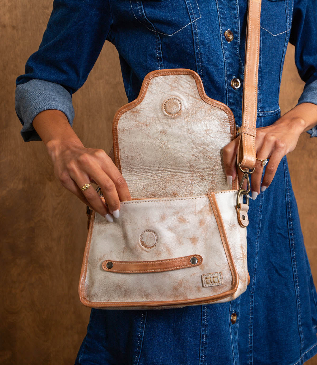 A person in a blue denim dress holds the open Ceremonious flap-top bag by Bed Stu, featuring light-colored leather with tan strap and trim, showing the inside pocket.