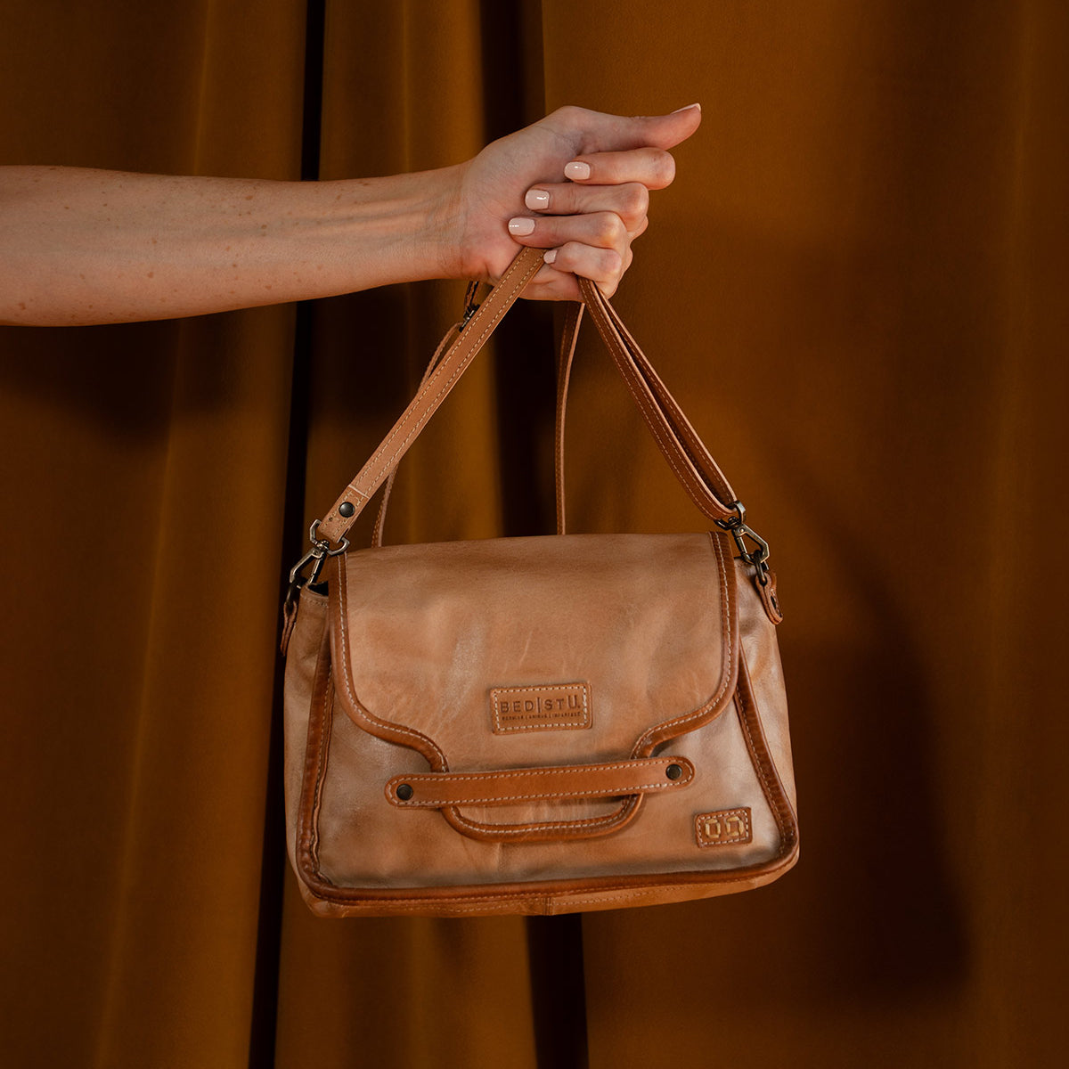 A hand holds the Ceremonious tan leather bag by Bed Stu, featuring a magnetic closure and embossed logo, set against a brown background.