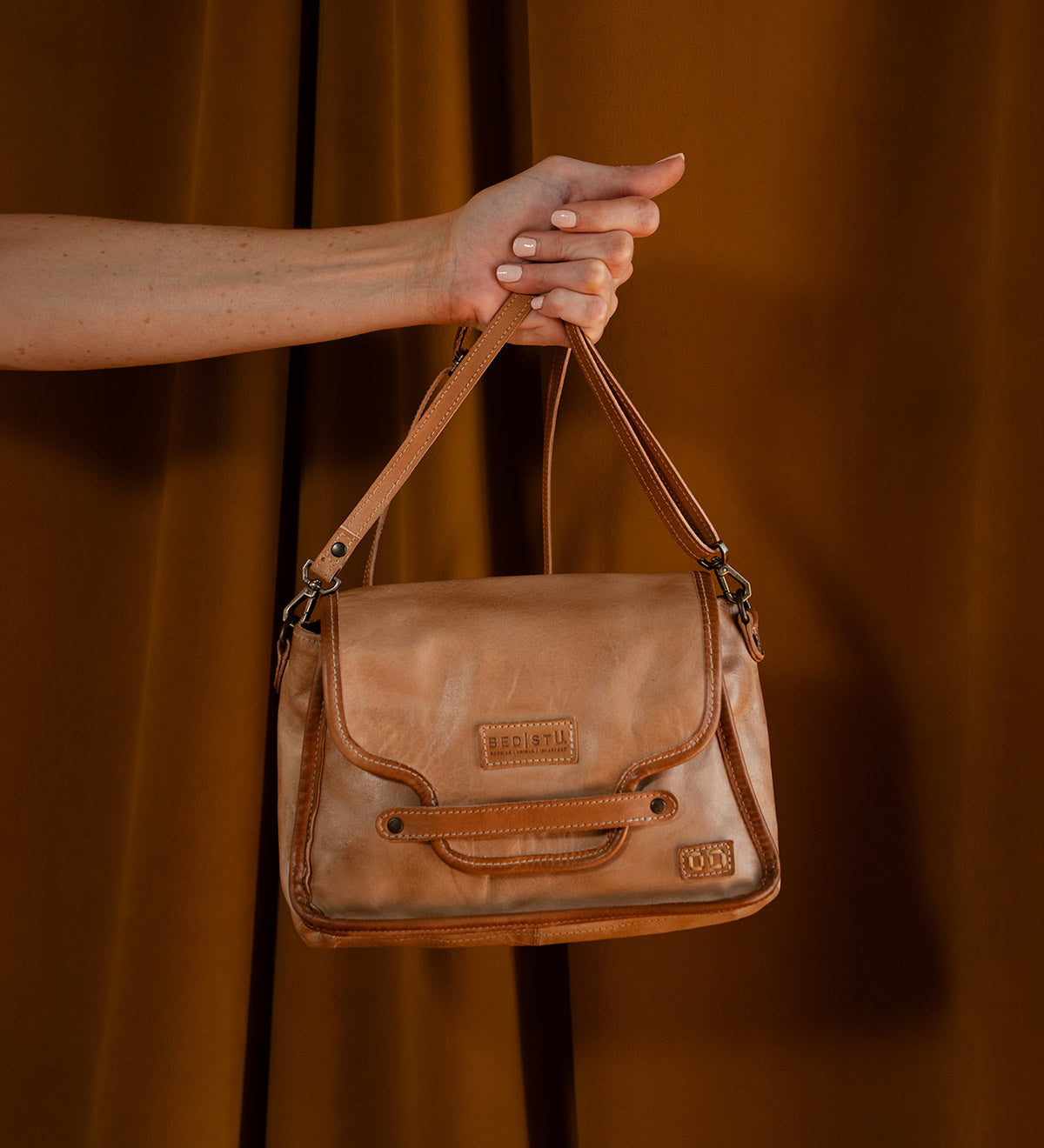 A hand holds the Ceremonious tan leather bag by Bed Stu, featuring a magnetic closure and embossed logo, set against a brown background.