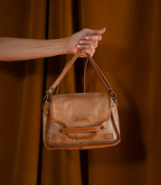 A hand holds the Ceremonious tan leather bag by Bed Stu, featuring a magnetic closure and embossed logo, set against a brown background.