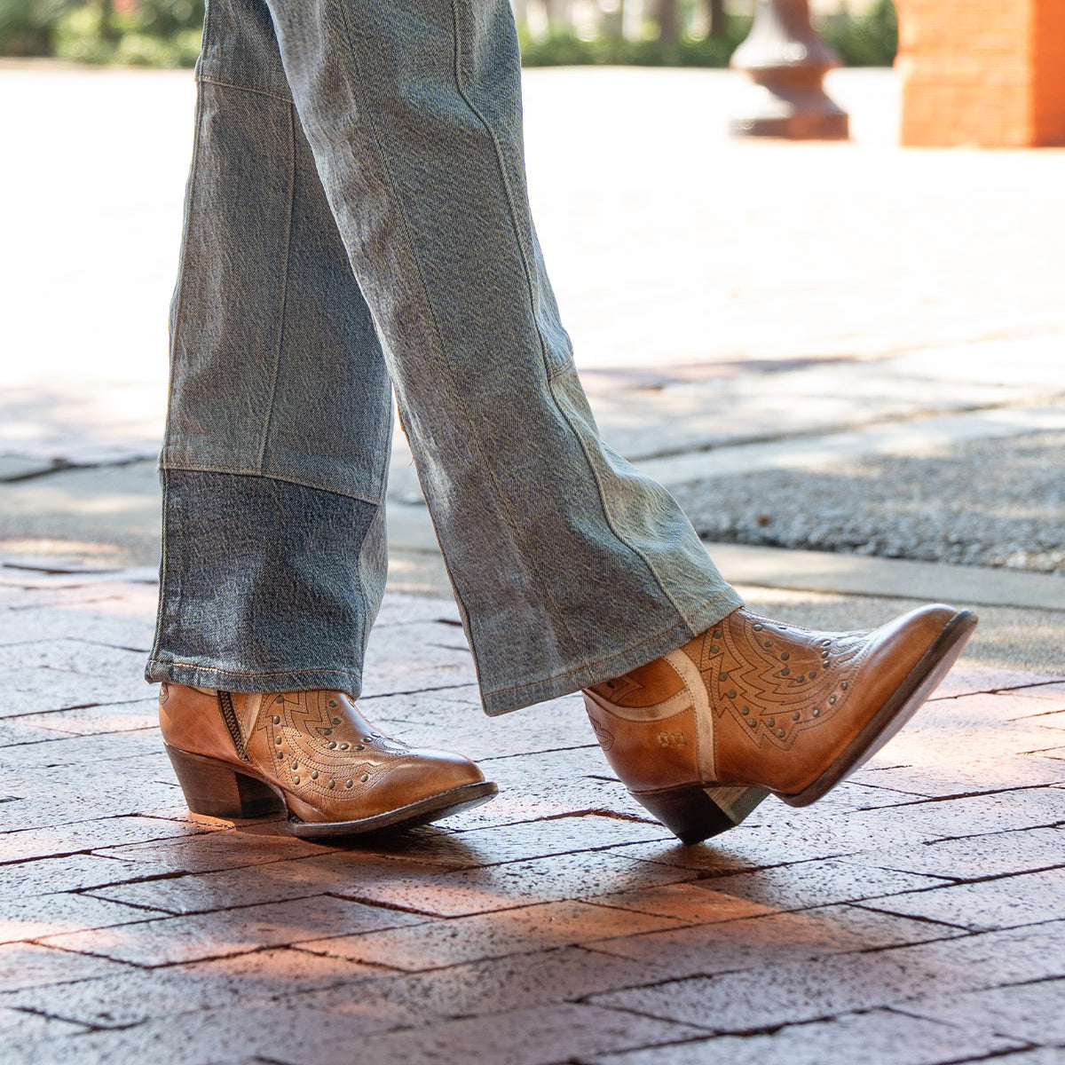 A person wearing Bed Stu's Chamise snip toe western boots with leather inlays and blue jeans walks on a brick sidewalk.