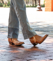 A person wearing Bed Stu's Chamise snip toe western boots with leather inlays and blue jeans walks on a brick sidewalk.
