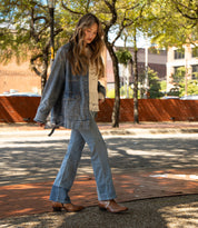 Wearing Bed Stu's Chamise denim jacket and jeans with mid-height boots, a person walks along a shaded, tree-lined sidewalk with buildings in the background.