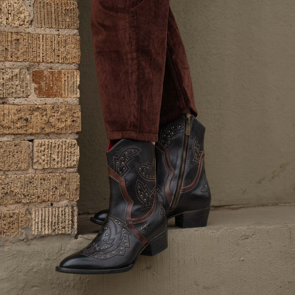 A person stands on a concrete ledge beside a brick wall, wearing Bed Stu's Chamise black western boots with a stacked heel and brown corduroy pants.