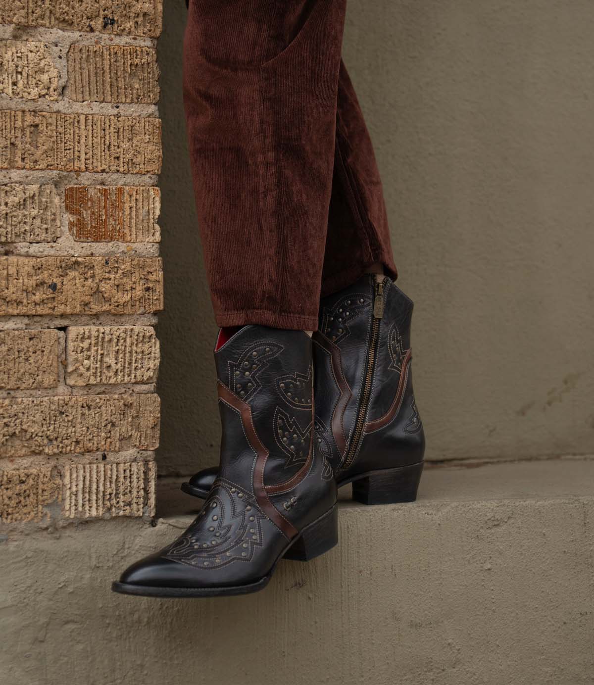 A person stands on a concrete ledge beside a brick wall, wearing Bed Stu's Chamise black western boots with a stacked heel and brown corduroy pants.