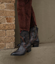 A person stands on a concrete ledge beside a brick wall, wearing Bed Stu's Chamise black western boots with a stacked heel and brown corduroy pants.