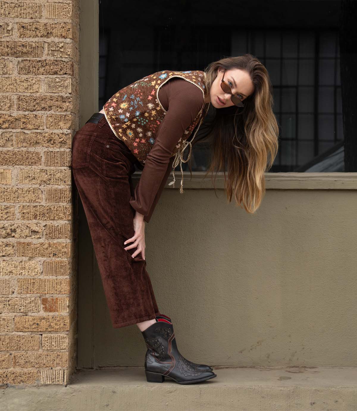 A woman wearing Bed Stu's Chamise floral vest, a brown shirt, brown pants, and stacked heel western boots leans against a beige wall and brick column, looking at the camera with sunglasses.