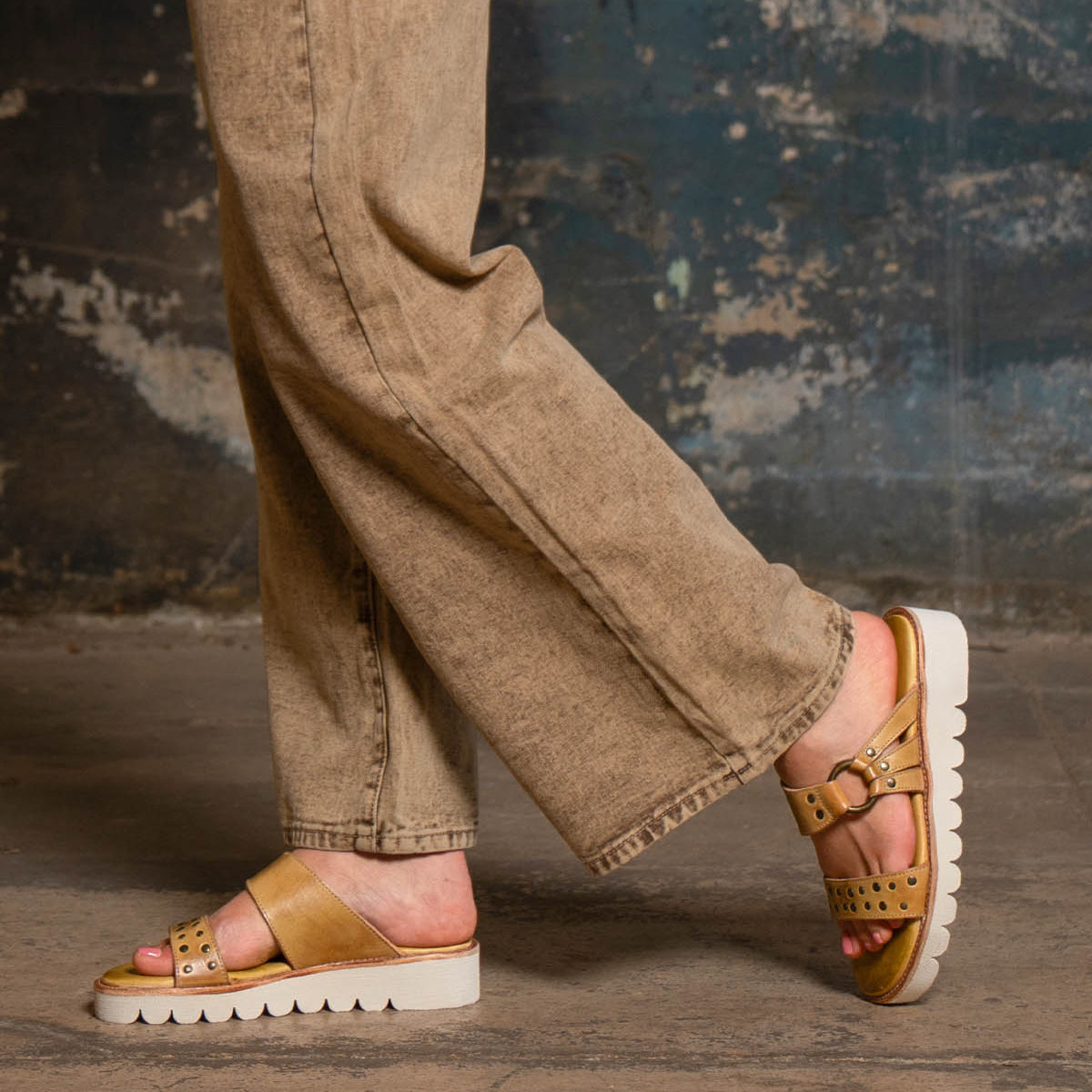 Wearing the Bed Stu Chow distressed leather sandals with buckle details and chunky rubber soles, paired with beige jeans, a person stands against a weathered, textured wall.