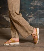 Wearing the Bed Stu Chow distressed leather sandals with buckle details and chunky rubber soles, paired with beige jeans, a person stands against a weathered, textured wall.