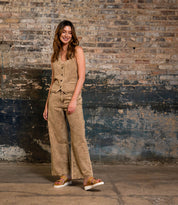 A woman stands indoors by a brick wall, wearing Bed Stu’s Chow sandals—statement footwear with a chunky rubber sole—paired with a tan sleeveless vest and matching wide-leg pants.